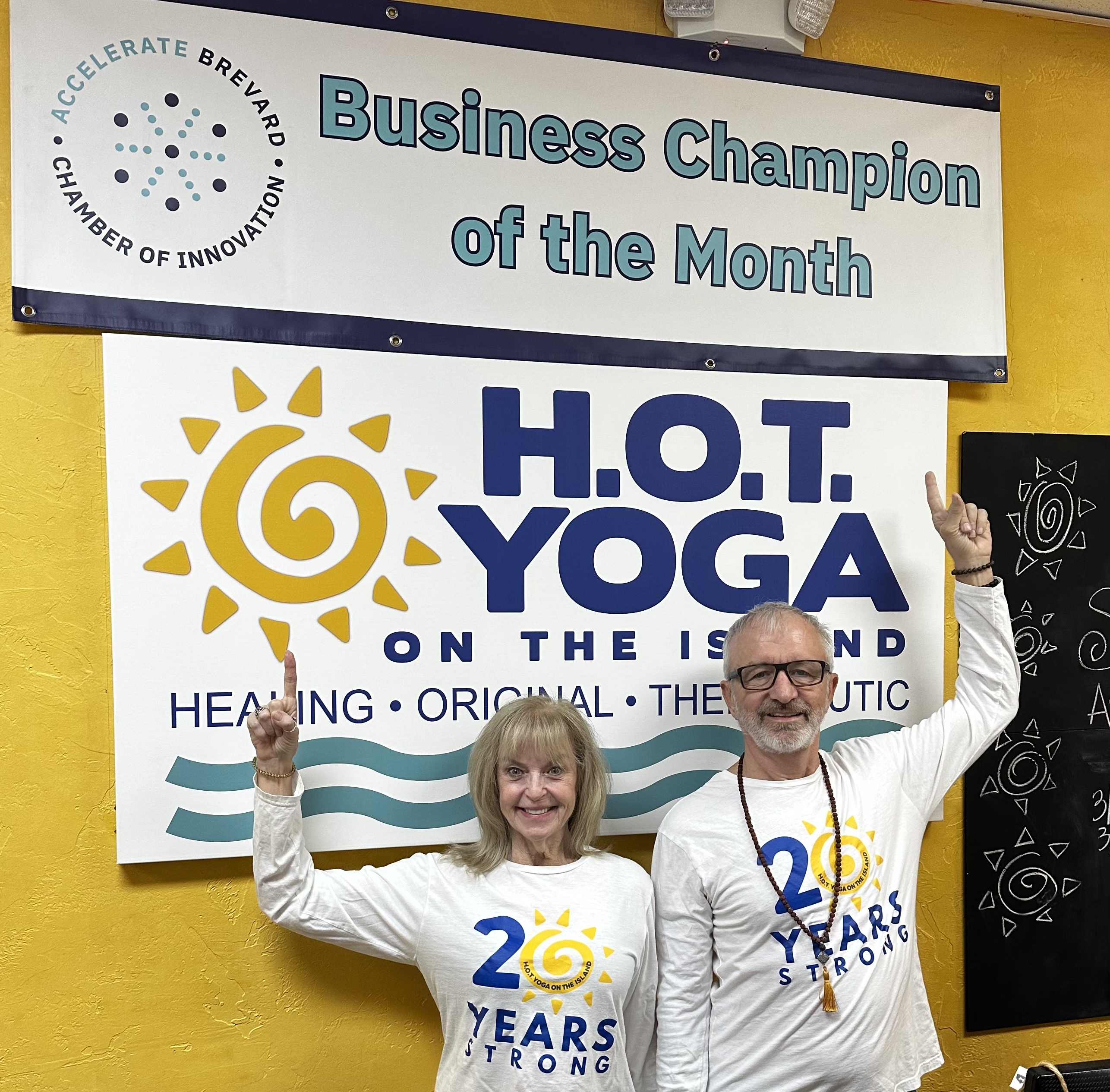 H.O.T. Yoga owners pointing at their Business Champion of the Month and 20 Years Strong banners
